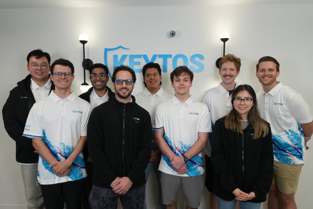 The Keytos team in the Boston office, standing together in front of the Keytos logo